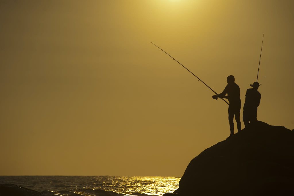 Silhouette Photo of Two Men Holding Fishing Rods Against Body of Water on Hill